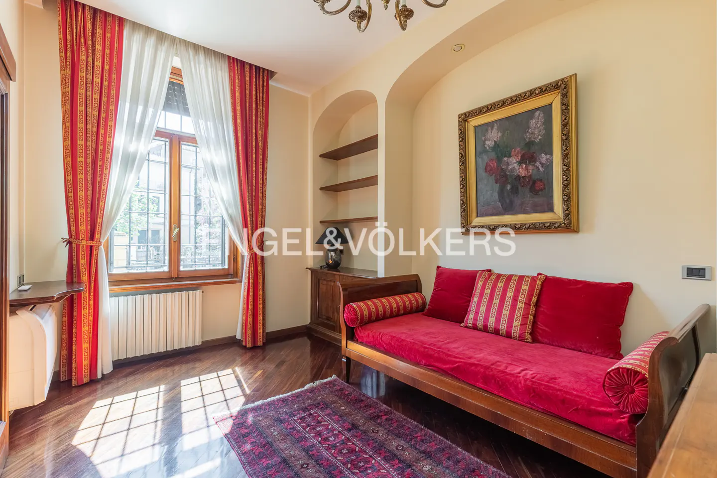 A cozy room with a red daybed, painting, and window with red curtains.