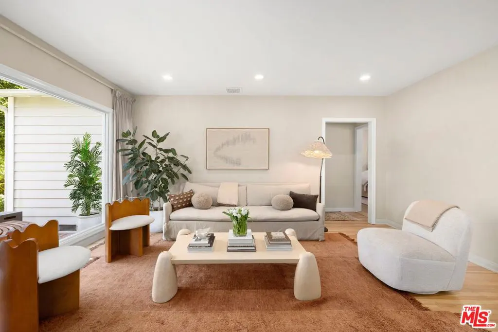 Bright living room with a beige sofa, unique coffee table, and two chairs on a brown rug. A large window and doorway let in natural light.