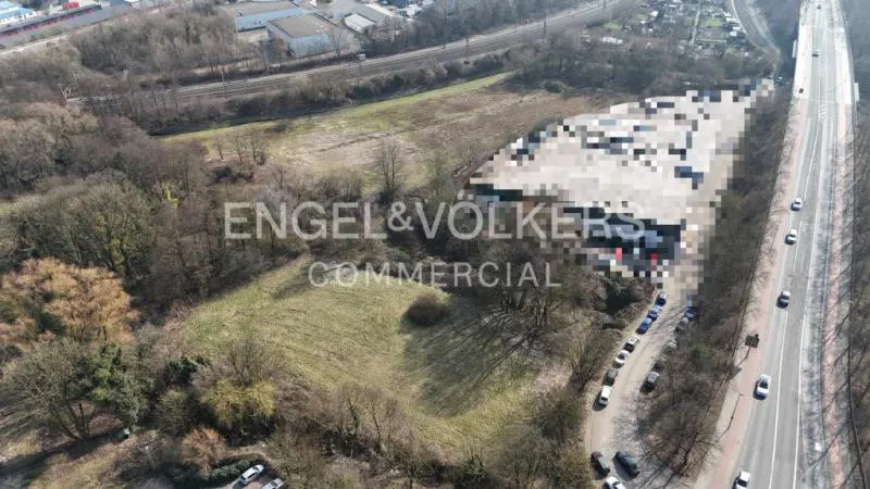 Aerial view of a commercial property with a blurred building, grassy land, trees, and a highway with cars. "Engel & Volkers Commercial" overlay.