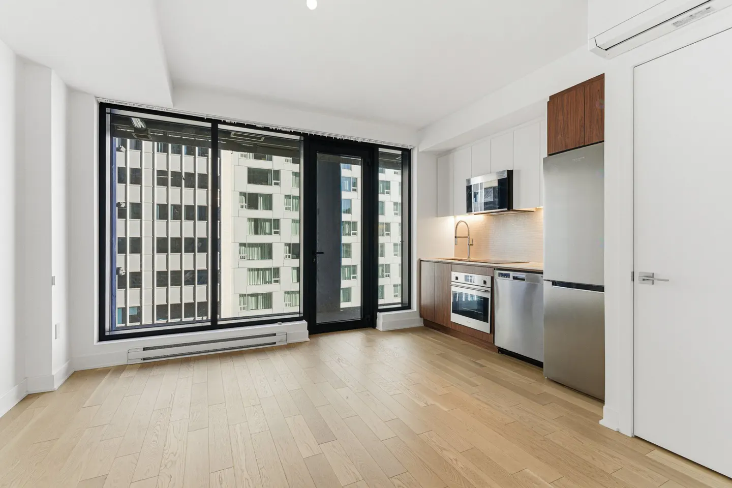 Modern kitchen with stainless steel appliances, wood cabinets, and light wood floors, next to a large window with city views.