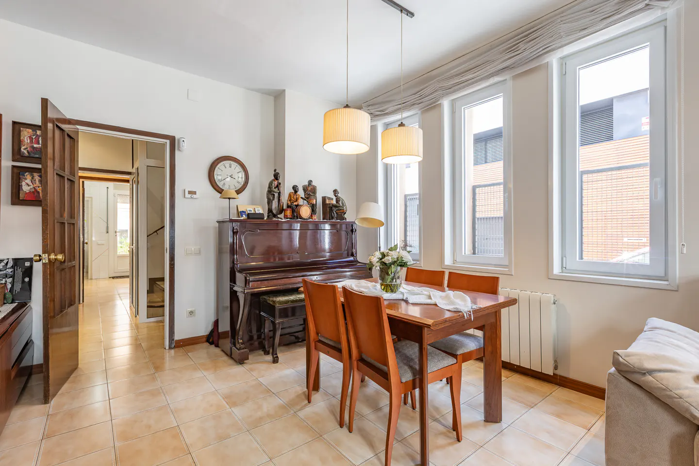 Dining room with a wooden table, four chairs, and an upright piano against a white wall under two pendant lights.
