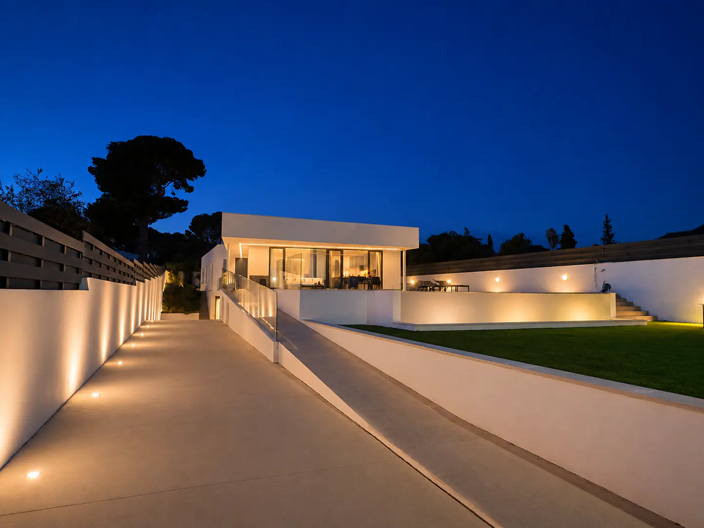 Modern white house at twilight with a lit driveway, ramp, and manicured lawn against a deep blue sky.