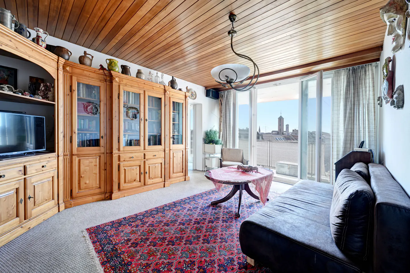 A living room with wood paneled ceiling, a large wooden cabinet, a blue sofa, and a red patterned rug. A balcony overlooks a city.