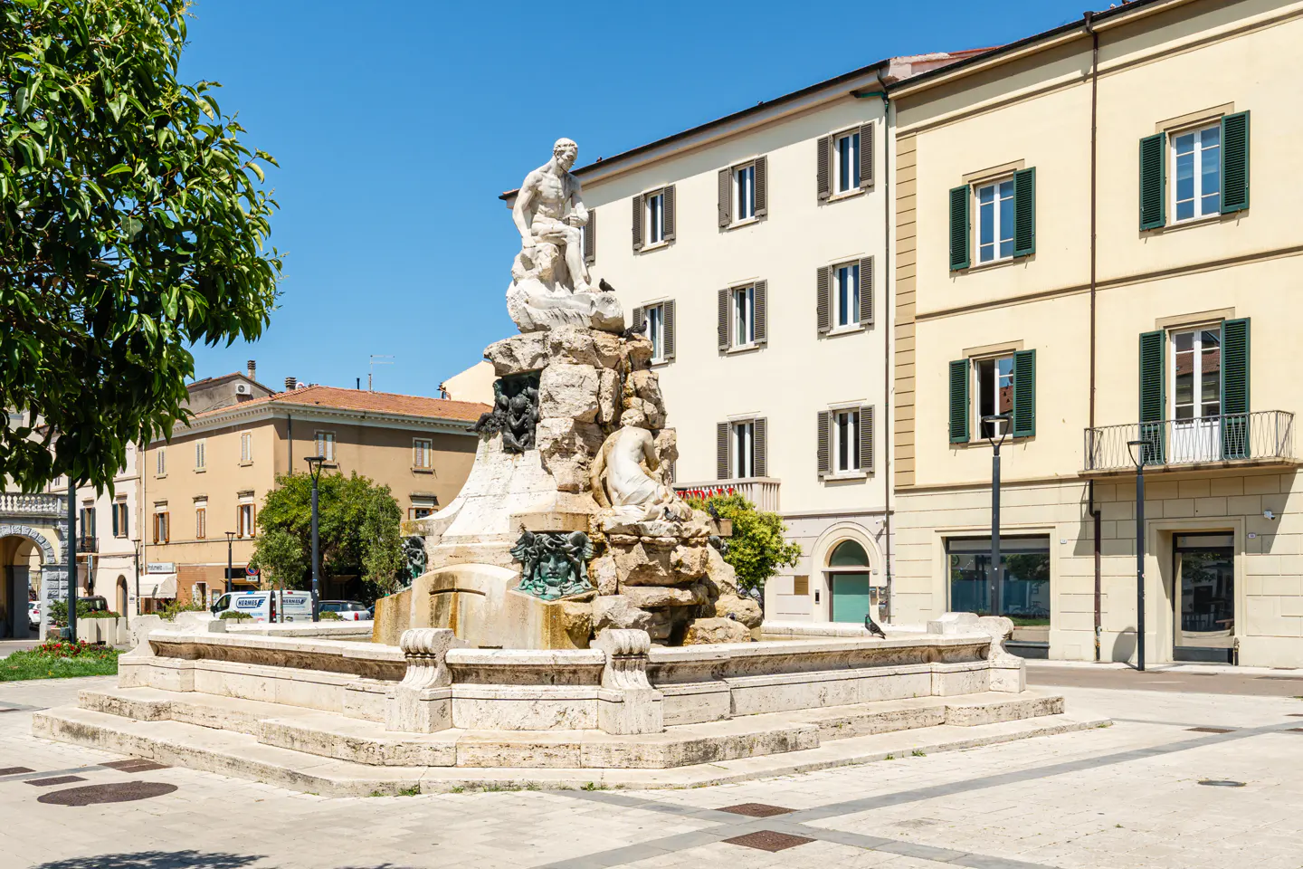 A stone fountain with classical statues stands in a sunny town square surrounded by multi-story buildings.