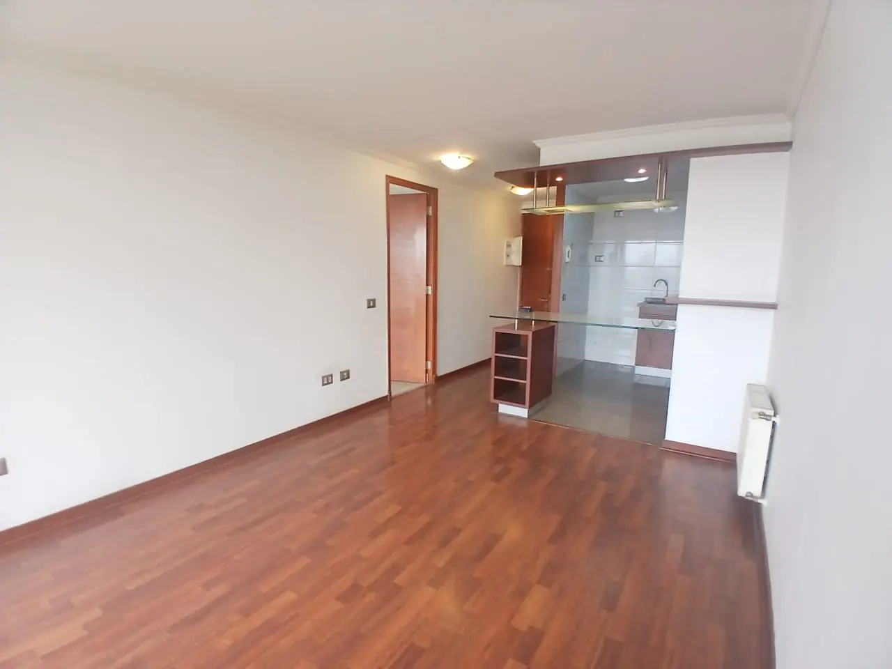 A bright, empty living room with hardwood floors and white walls. A doorway and kitchen bar area are visible.