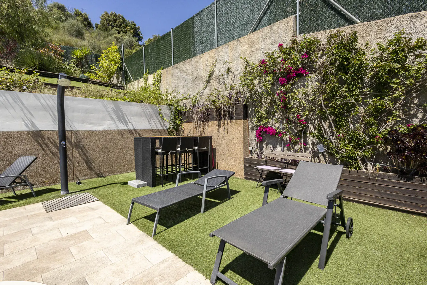 Outdoor patio with lounge chairs, a high-top table with stools, and a shower. A wall with climbing plants and pink flowers is in the background.