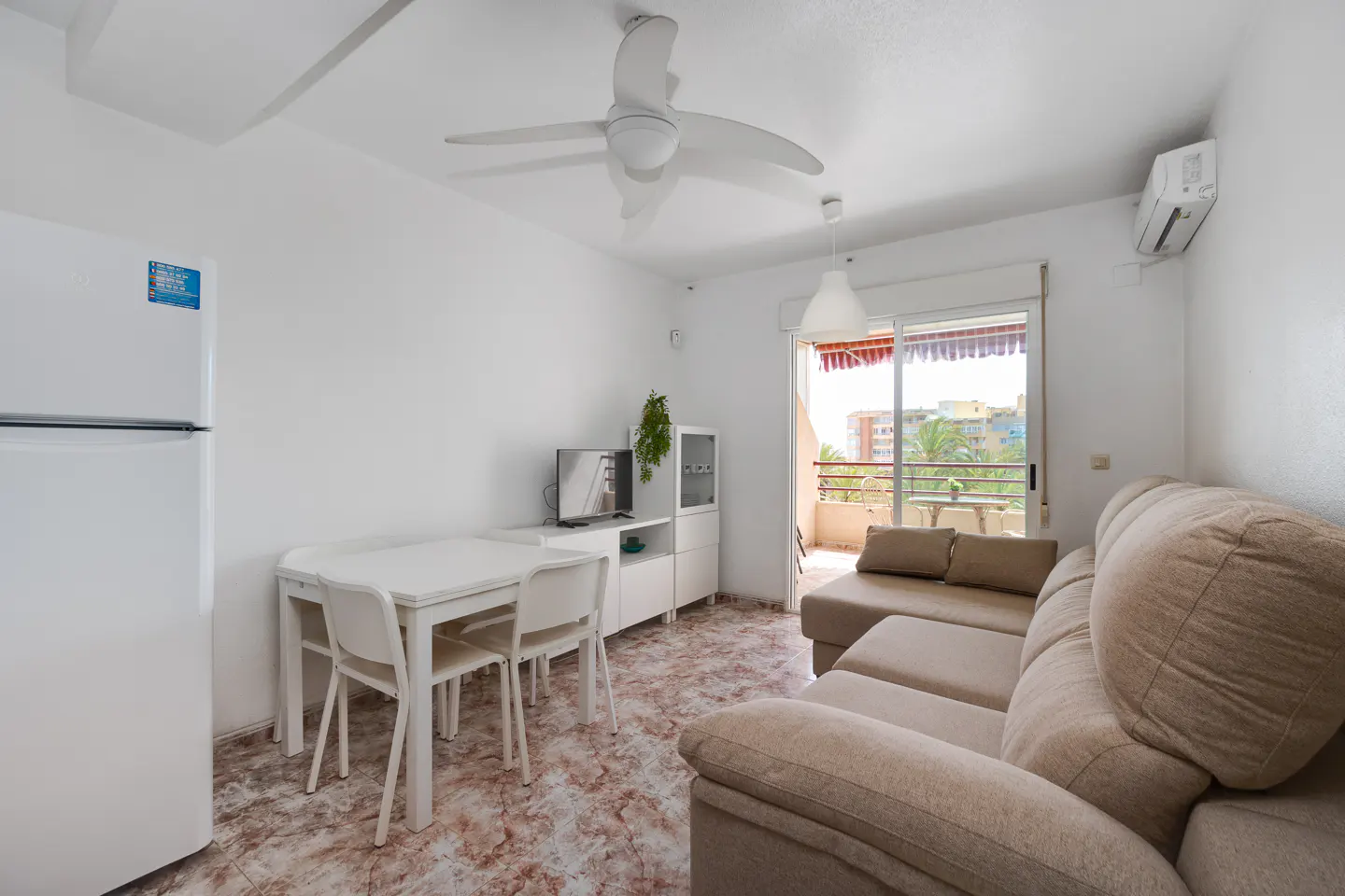 Bright living space with white walls, tile floor, and beige sectional sofa. A white dining set sits near a white refrigerator. Balcony with palm tree view.