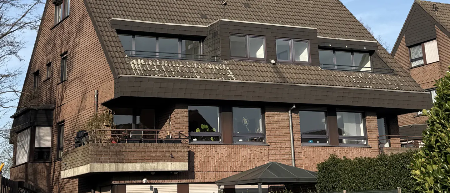 A three-story brick building with a brown tiled roof and dark-framed windows. Balconies are visible on the lower floors.