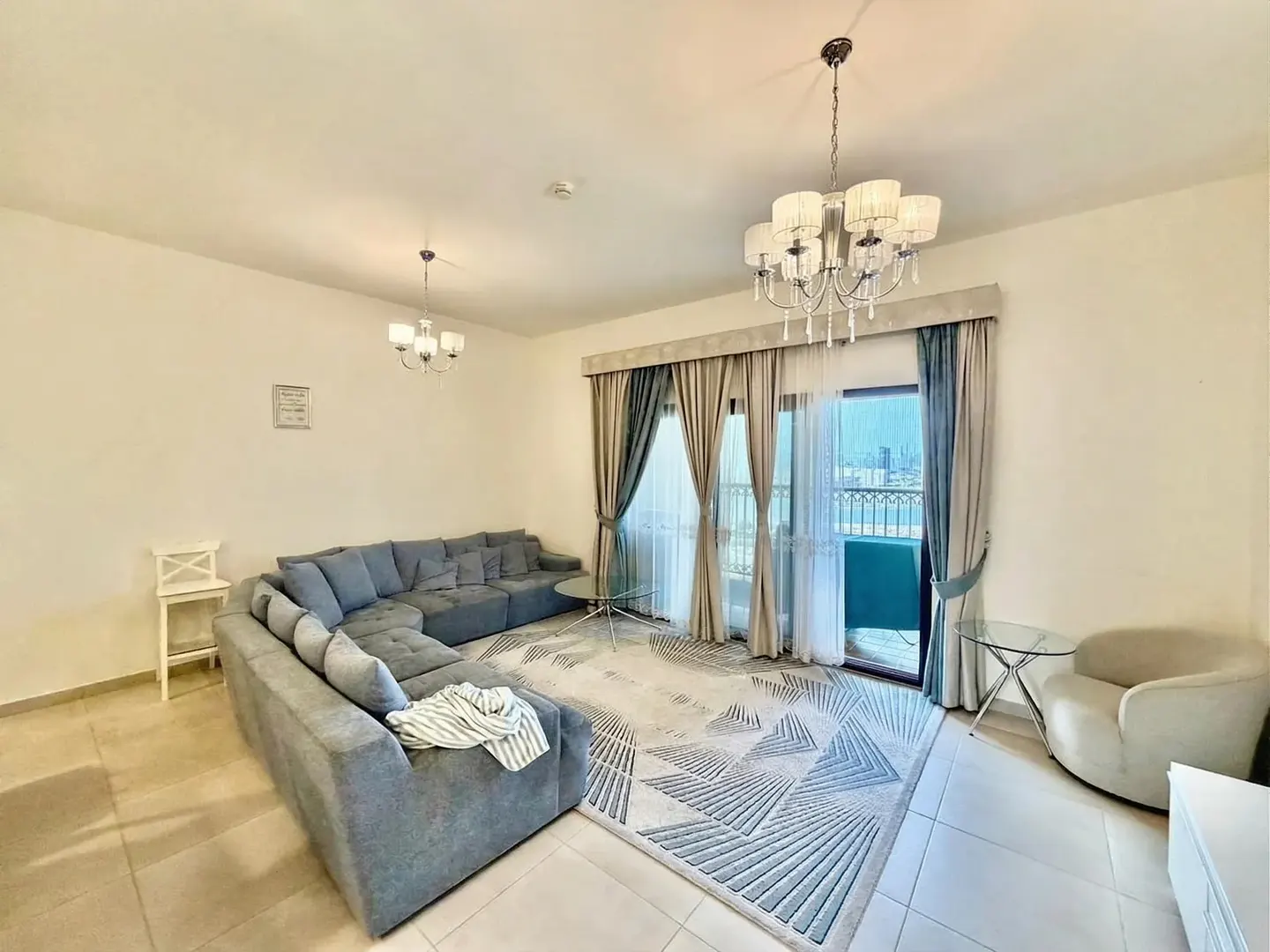 Living room with gray sectional sofa, geometric rug, and balcony with city view. Chandelier and neutral walls.