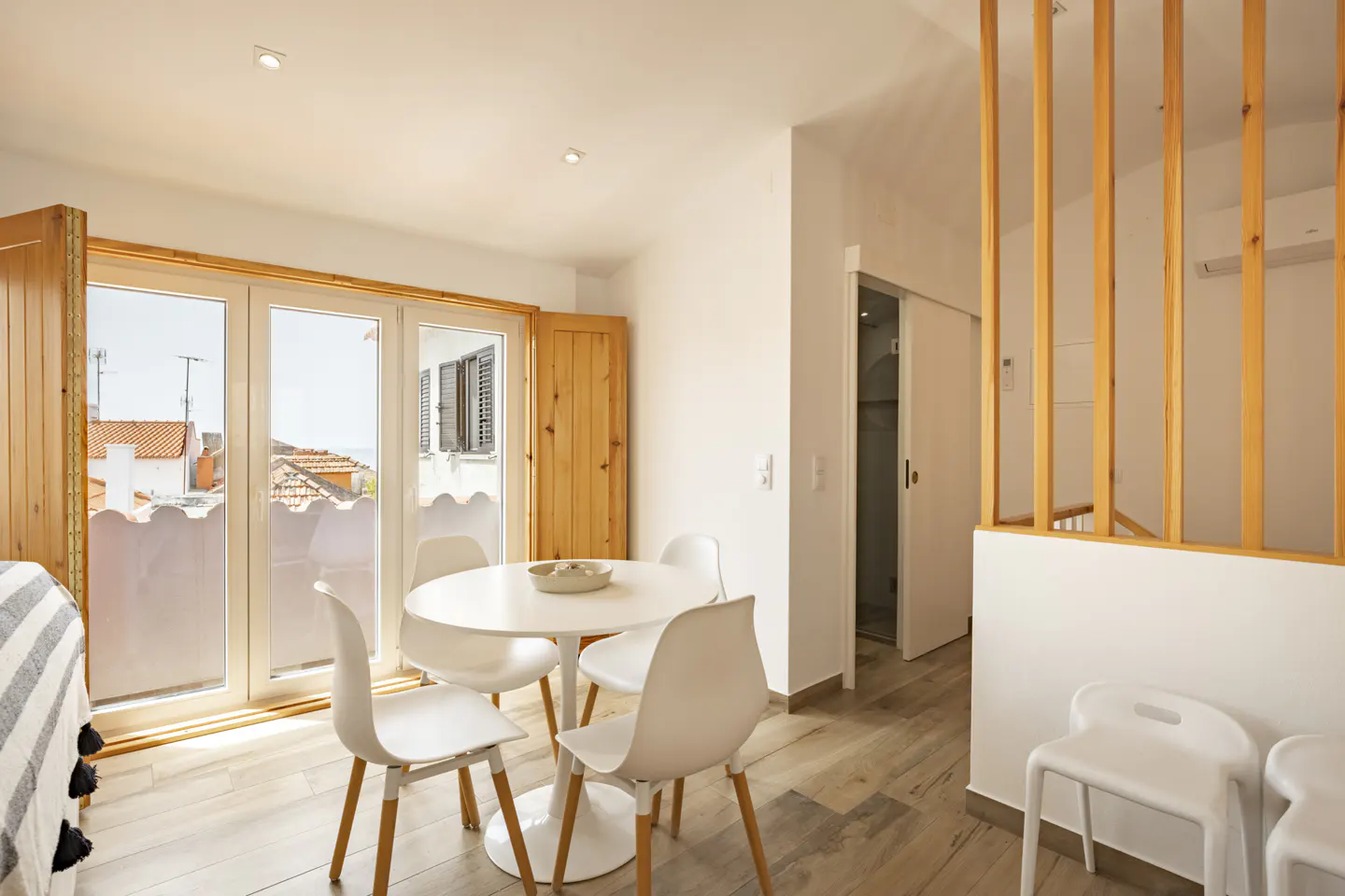 Bright apartment interior with a round white table and four chairs near open doors to a balcony with a view of rooftops.