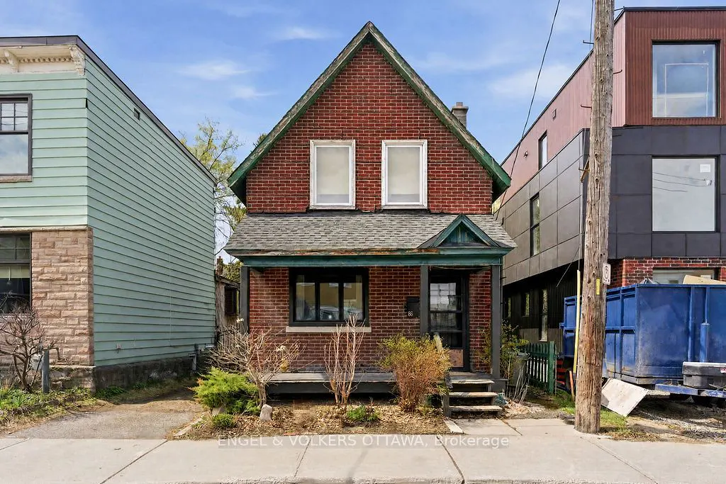 Two-story red brick house with a gray roof and green trim, flanked by other buildings under a blue sky.