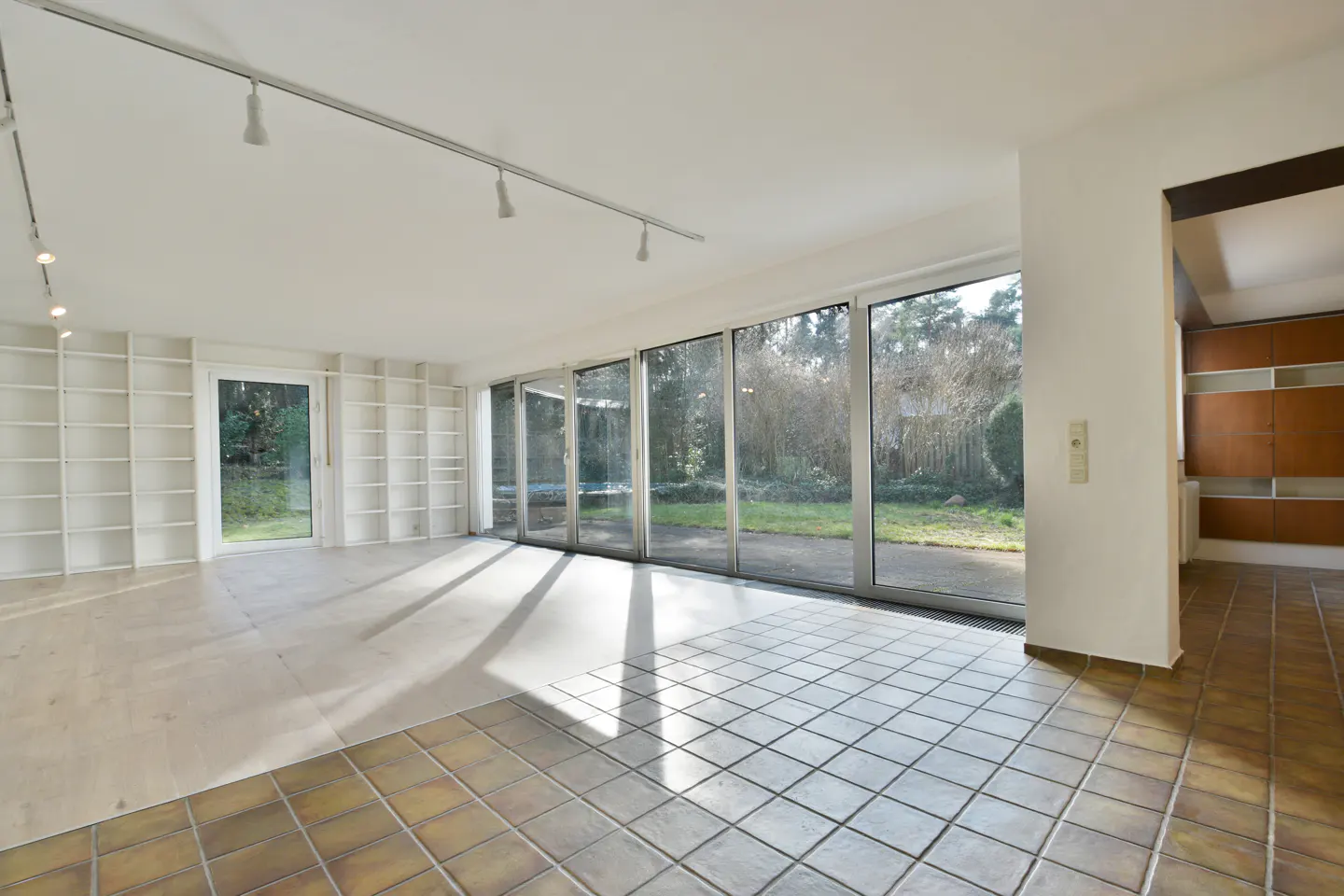 Bright, empty room with white walls, track lighting, and large windows overlooking a green yard. Built-in white shelves and brown tiled floor.