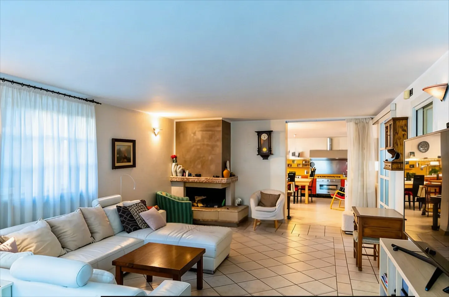 Bright living room with a white sectional sofa, dark wood coffee table, and a fireplace. A kitchen is visible in the background.