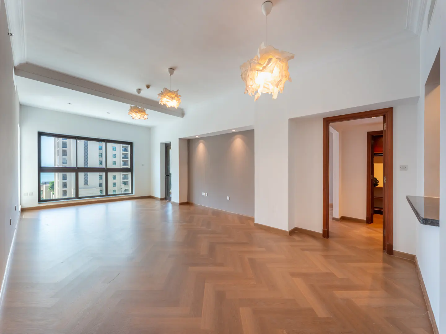 Bright, empty apartment with herringbone wood floors, white walls, and modern light fixtures. A large window shows a city view.