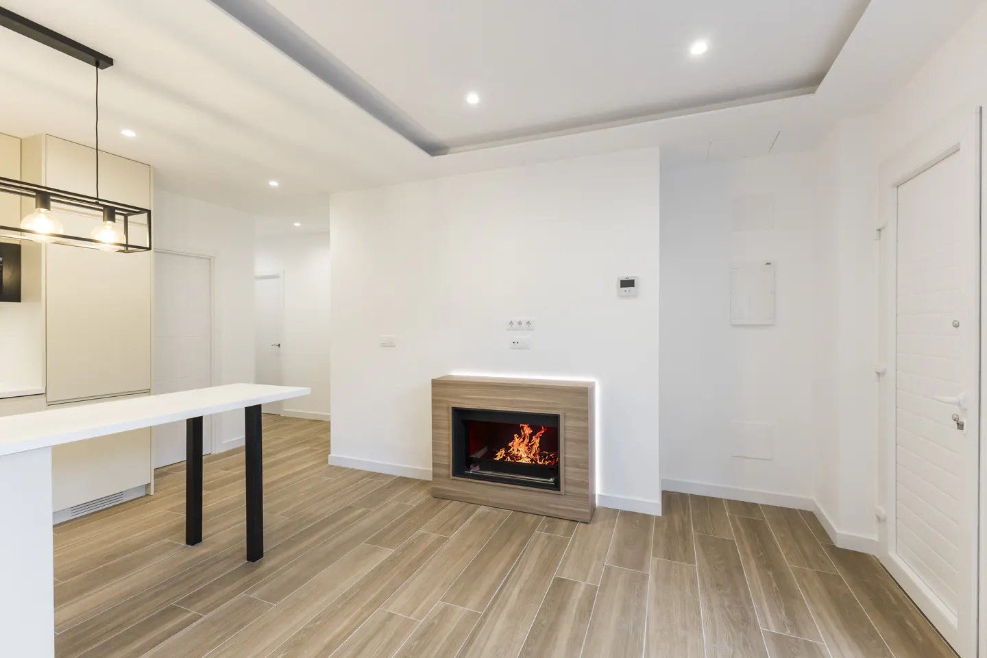 Bright, modern living room with a fireplace, white walls, and wood-look tile flooring. A white kitchen island is visible.