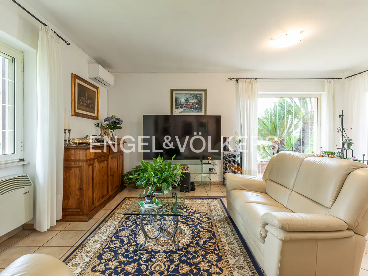 Living room with a cream sofa, blue patterned rug, glass coffee table, and a large TV. White walls and curtains.