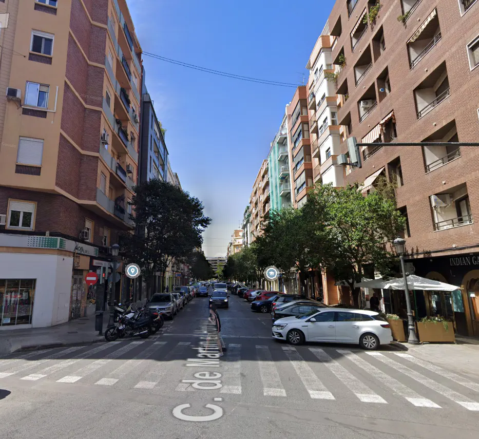 Street view of C. de Martí in Valencia, Spain, with parked cars, trees, and brick buildings under a blue sky.