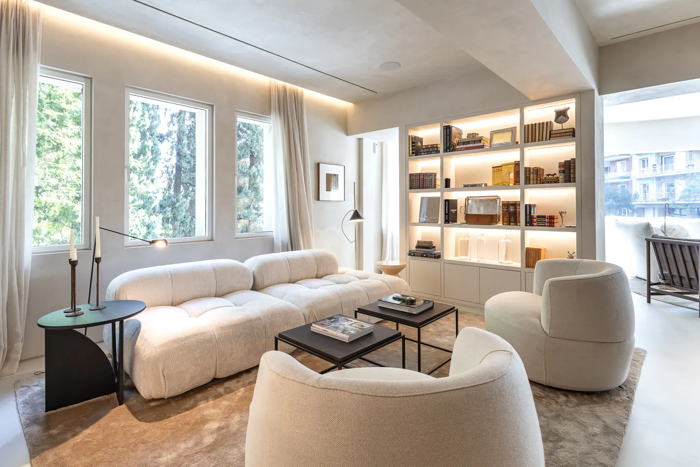 Bright living room with a white sofa, two chairs, and black tables on a brown rug. A bookcase with books and decor is lit from behind. Windows show trees.