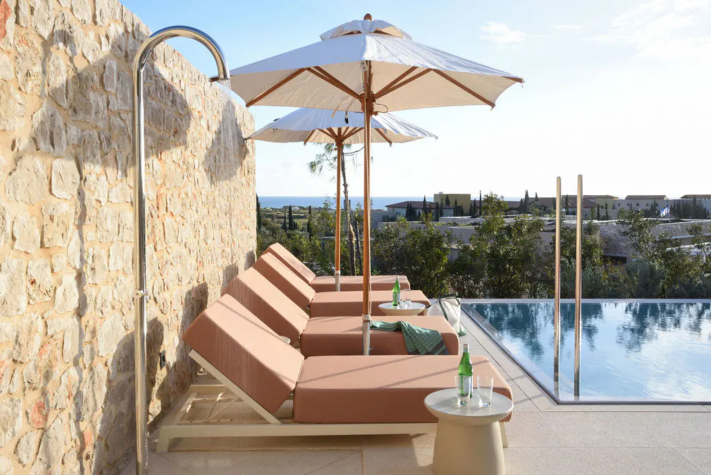 Outdoor lounge area with tan chaise lounges, white umbrellas, and a pool overlooking the ocean. A stone wall and shower are on the left.