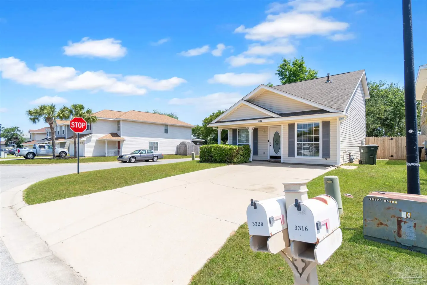 A beige single-story house with a gray roof, white trim, and a concrete driveway on a sunny day. Two white mailboxes are in the foreground.
