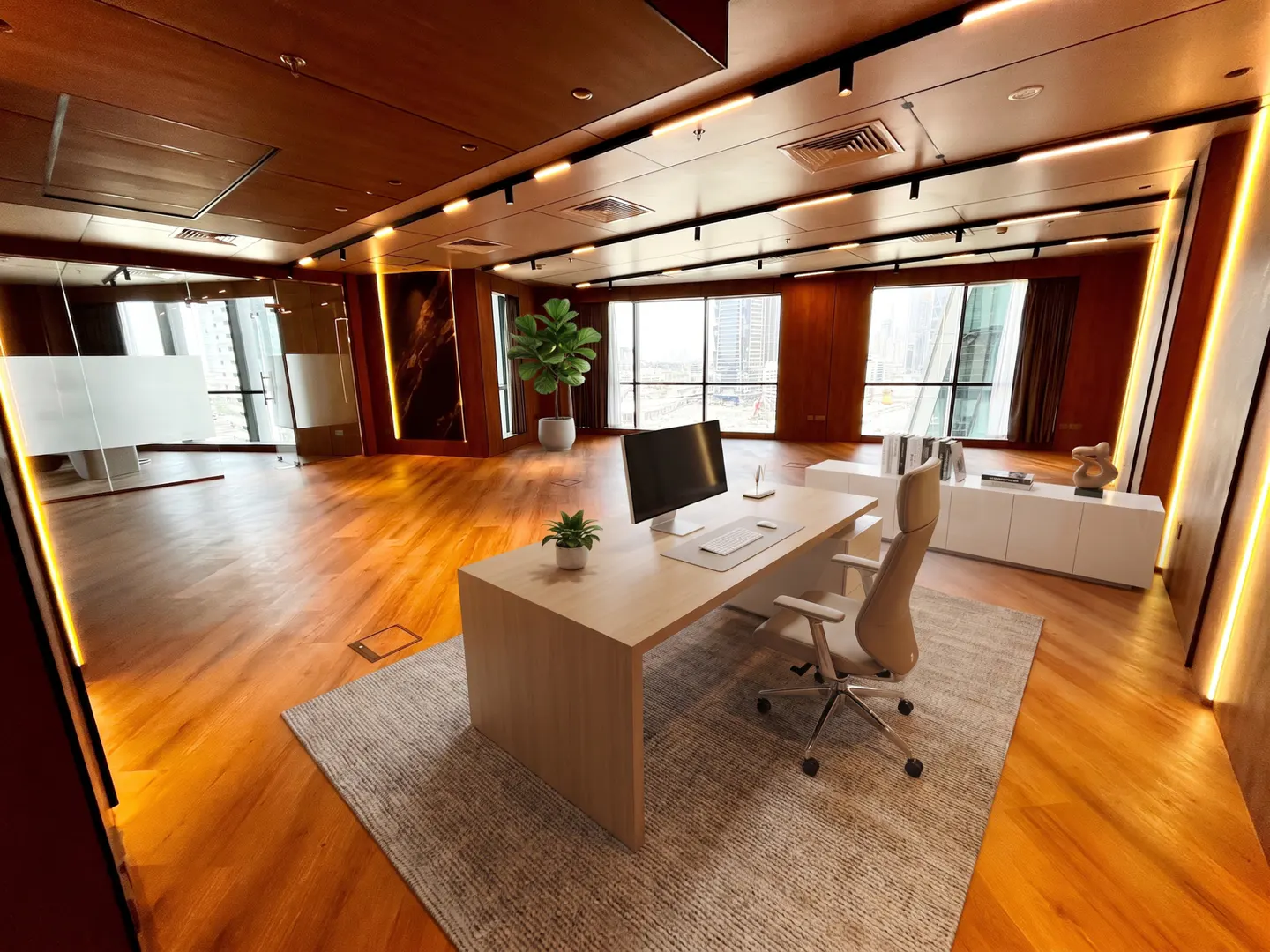 Modern office space with wood floors, large windows, and a desk with a computer and chair. A plant and white cabinets add to the decor.