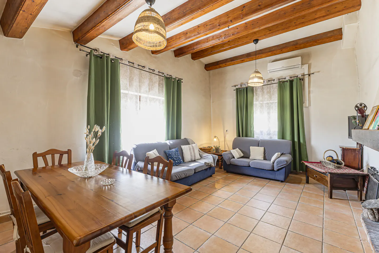 Living room with wood beams, tile floor, and two blue sofas. A wood table and chairs are in the foreground. Green curtains frame the windows.