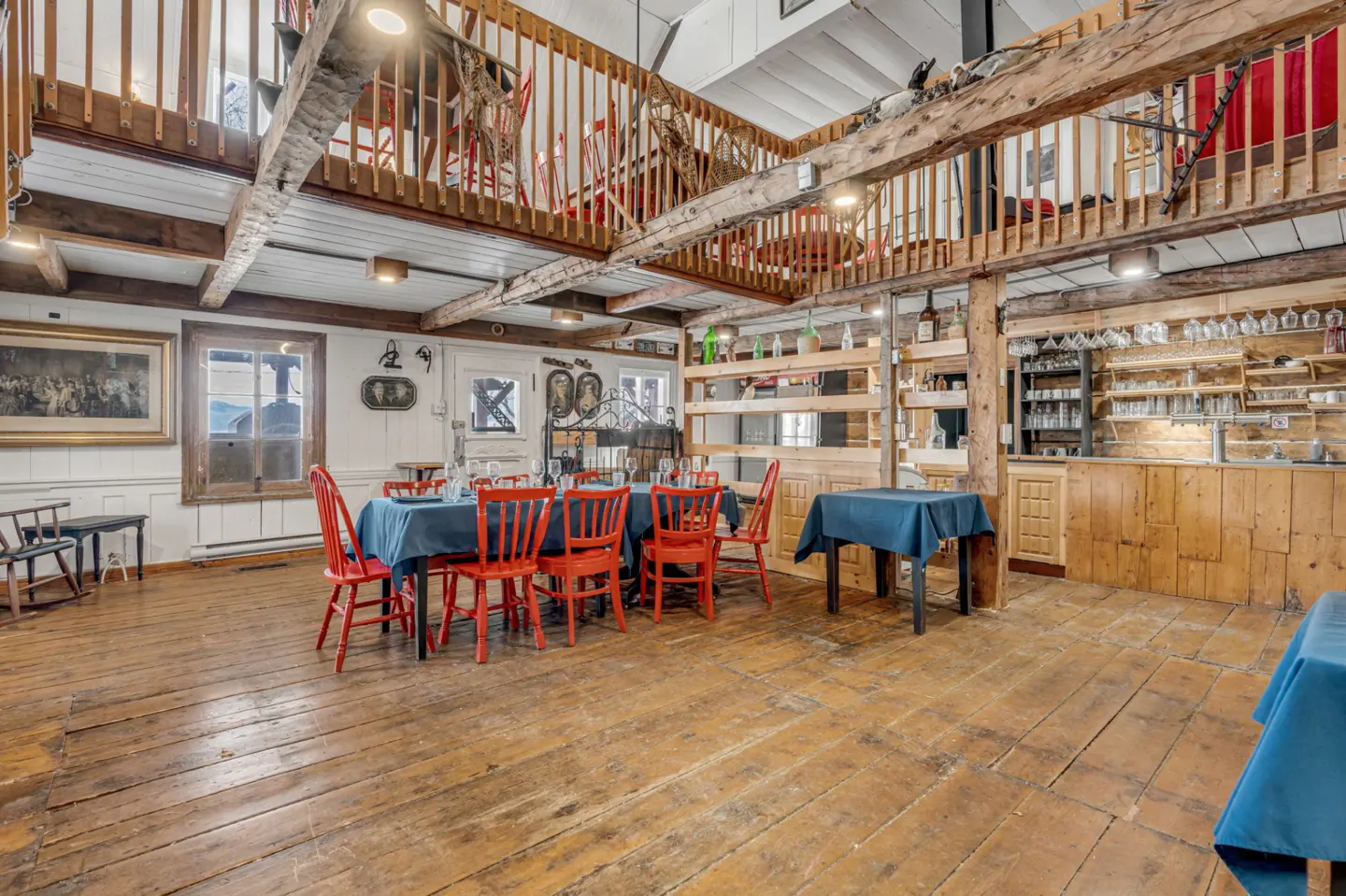 Rustic dining room with wood floors, exposed beams, and a loft. Red chairs surround tables with blue cloths. A wooden bar is on the right.