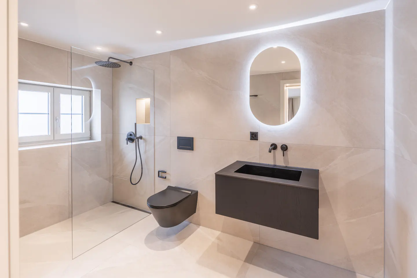 Modern bathroom with beige tile, black toilet and sink, and an oval LED mirror. Shower with glass door and window.