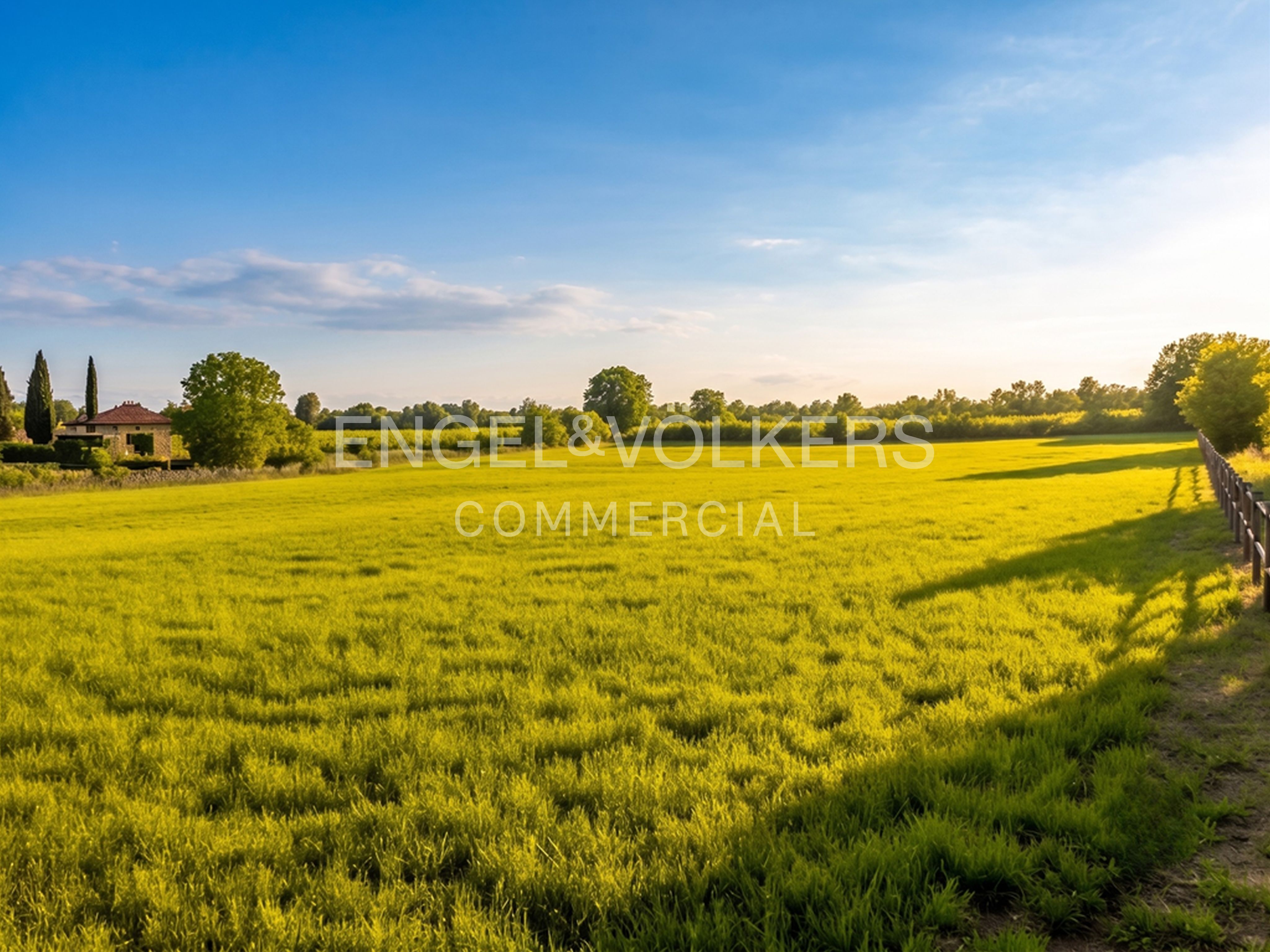 A vast, sunlit green meadow under a clear blue sky, with a distant house, trees, and a wooden fence along the right side.
