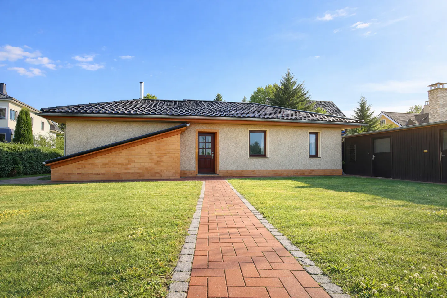 A single-story beige house with a dark roof and a brick walkway leading to the front door. Green lawn and blue sky.