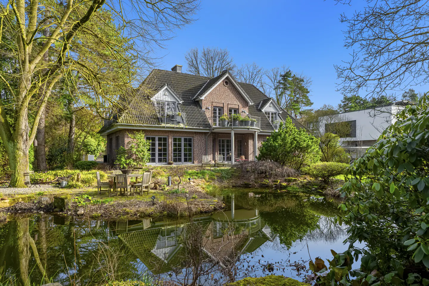 Brick house with balconies reflected in a pond, surrounded by trees and greenery under a blue sky.
