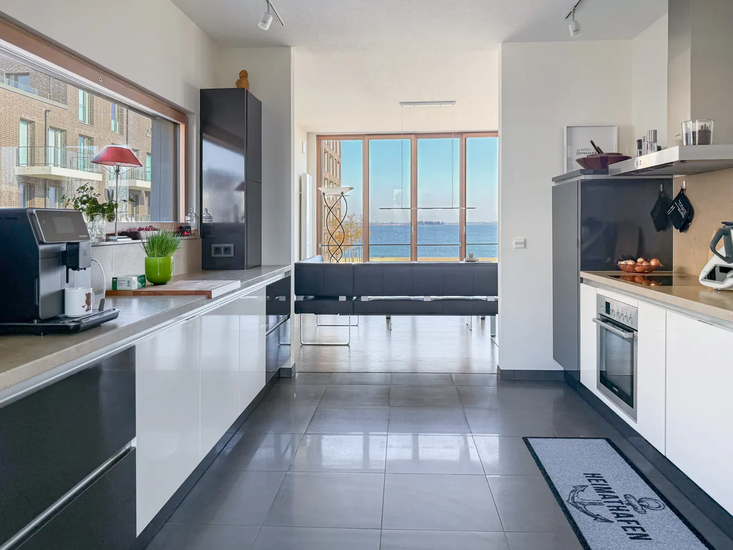 A modern kitchen with white cabinets, gray tile floor, and a view of the ocean through large windows. A black coffee maker sits on the counter.