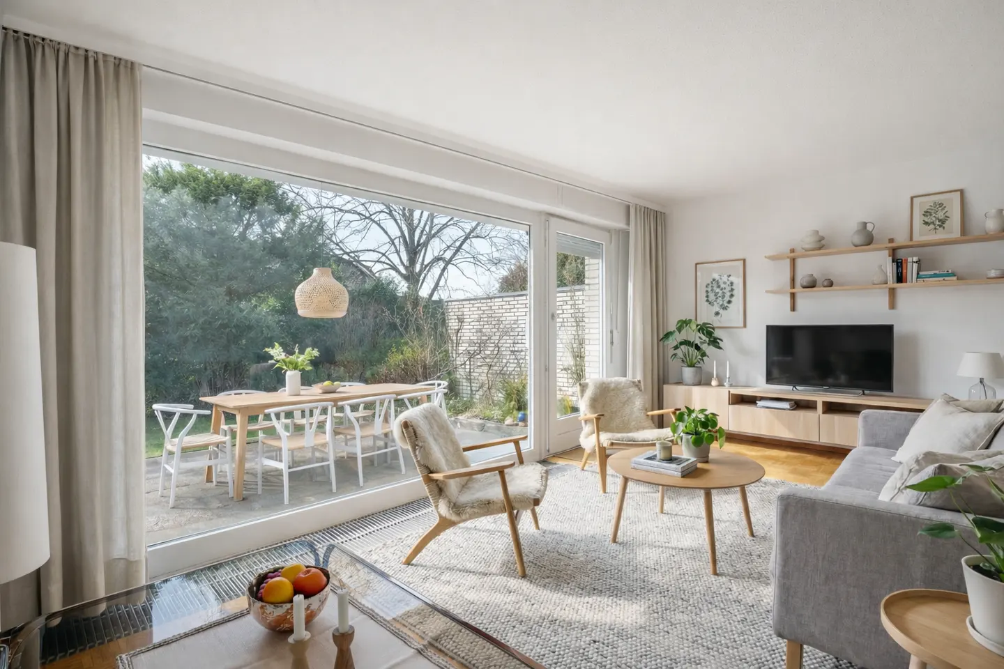 Bright living room with a gray sofa, wood furniture, and a large window overlooking a patio with a dining table.