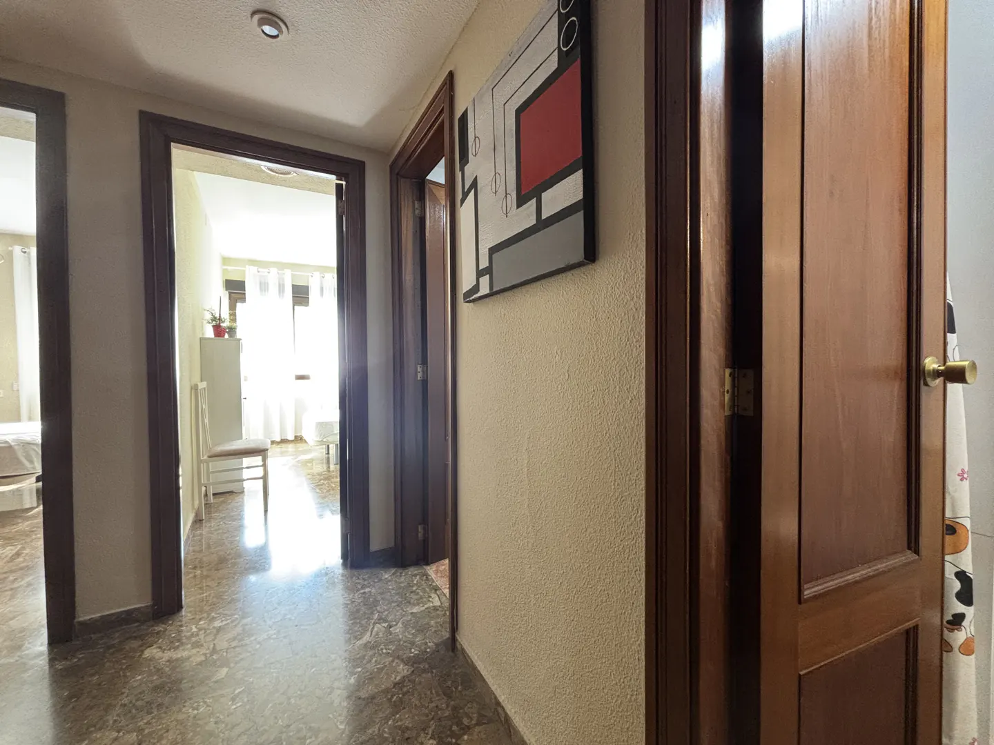 Hallway with marble floors, beige walls, and dark wood trim. Abstract art hangs on the wall. Open doorways lead to sunlit rooms.