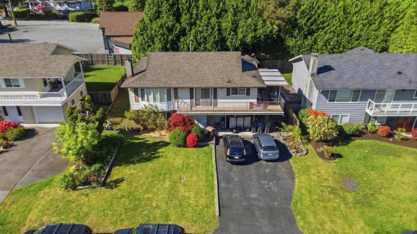 Aerial view of a two-story house with a brown roof, a balcony, and two cars parked in the driveway. Green lawn and trees surround the house.