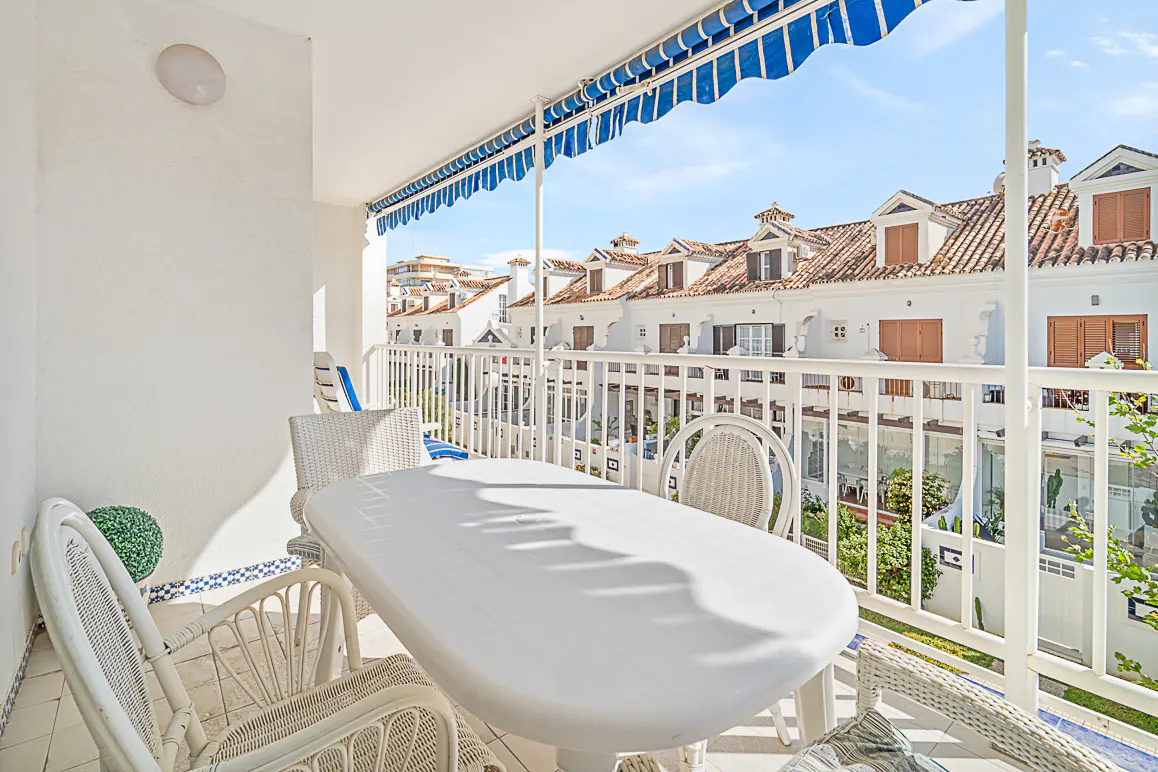 Balcony with white wicker furniture, table, and blue striped awning. White buildings with terracotta roofs in the background.