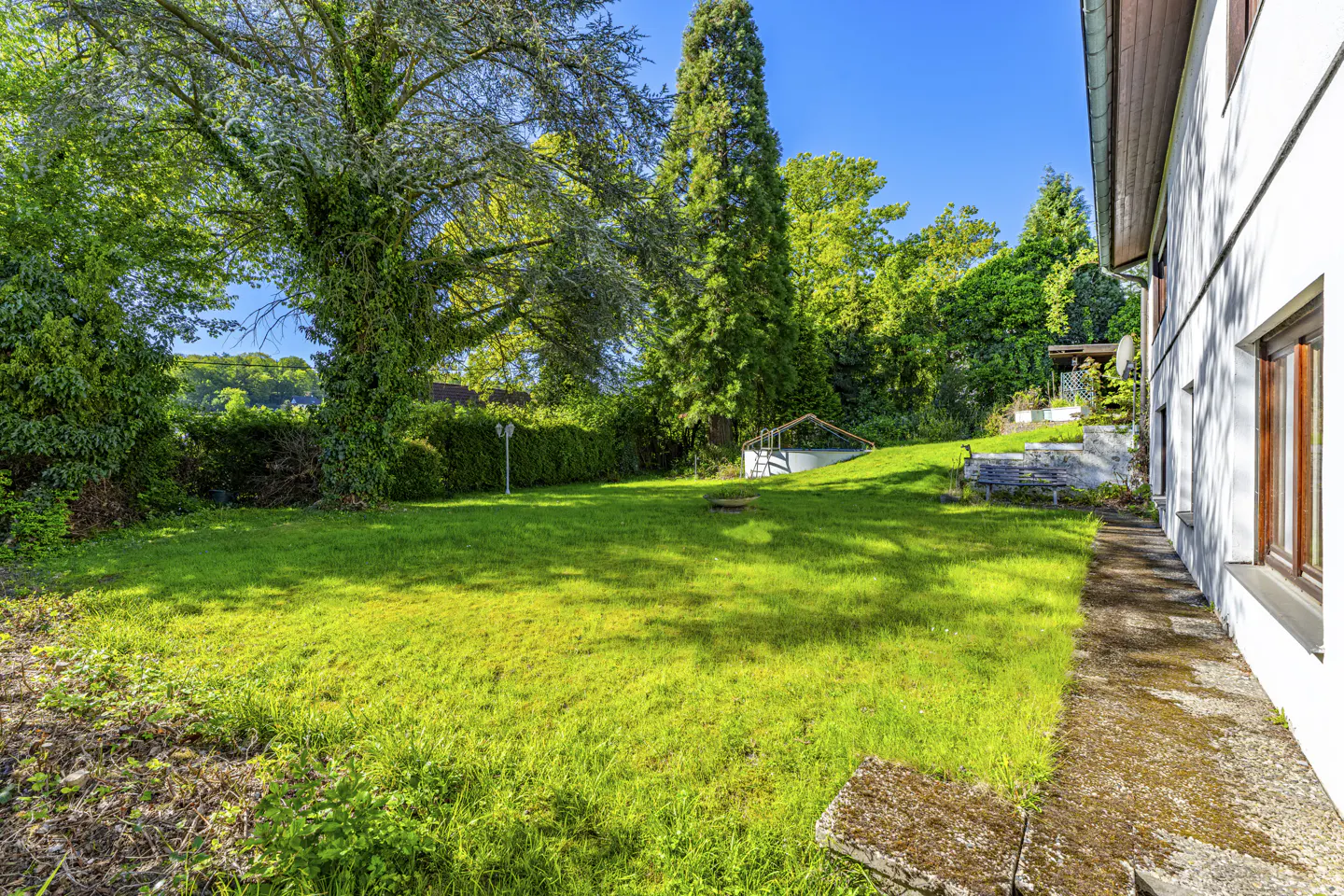 A lush green lawn with trees and a white house on the right. A stone path leads along the house. A bench and small structure are visible in the background.
