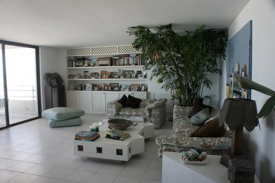 Bright living room with white tile floors, patterned sofas, and a white bookshelf filled with books and photos. A large plant adds a touch of nature.