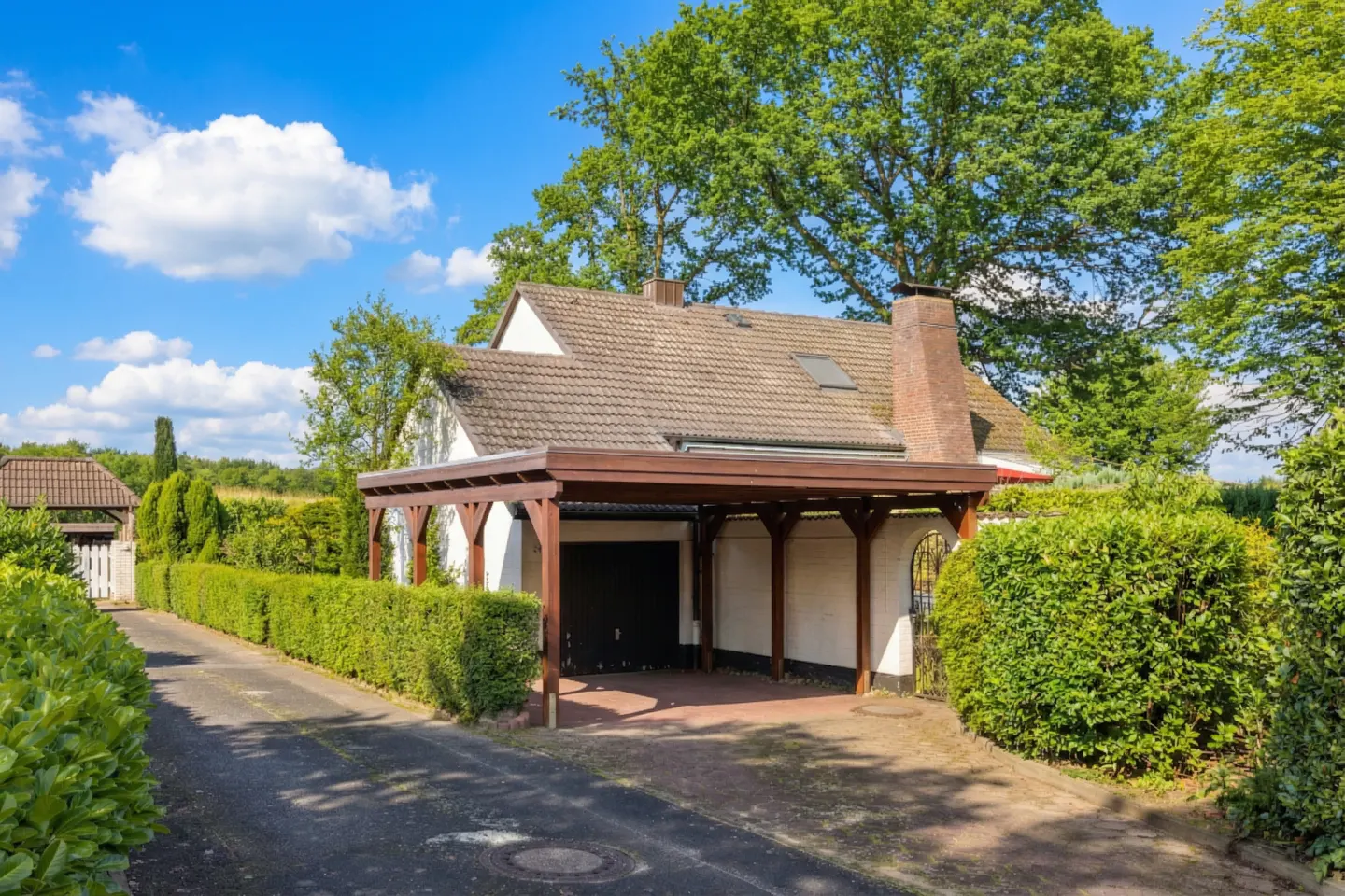 Exterior of a white house with a brown carport, tile roof, and brick chimney under a blue sky with clouds.
