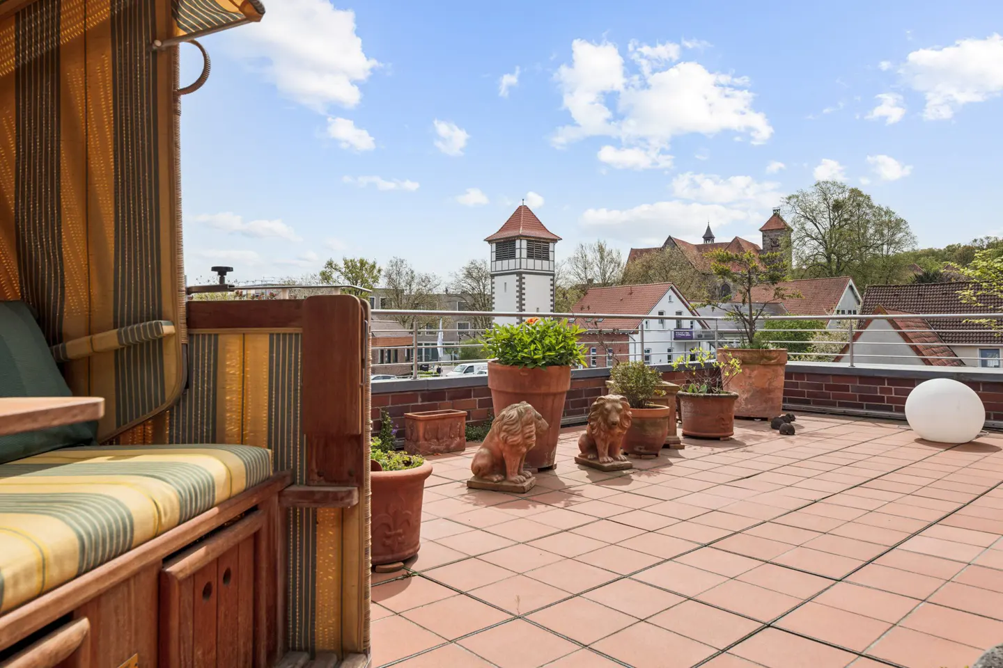 Rooftop terrace with a striped beach chair, terracotta tiles, potted plants, lion statues, and a view of buildings under a blue sky.