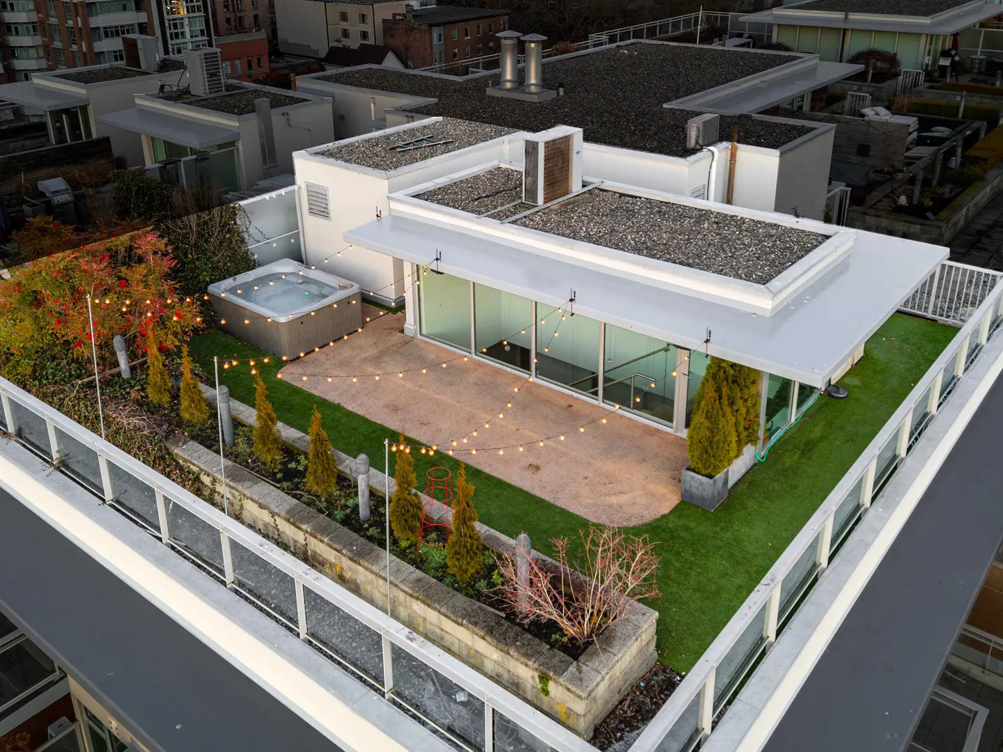 Rooftop patio with hot tub, string lights, and greenery. Modern white building with glass walls. Cityscape background.