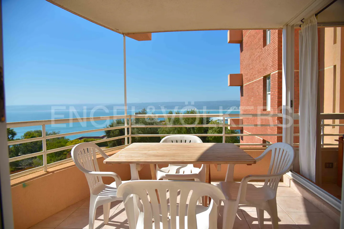 Balcony with a wooden table and white plastic chairs overlooking the ocean and a red brick building.