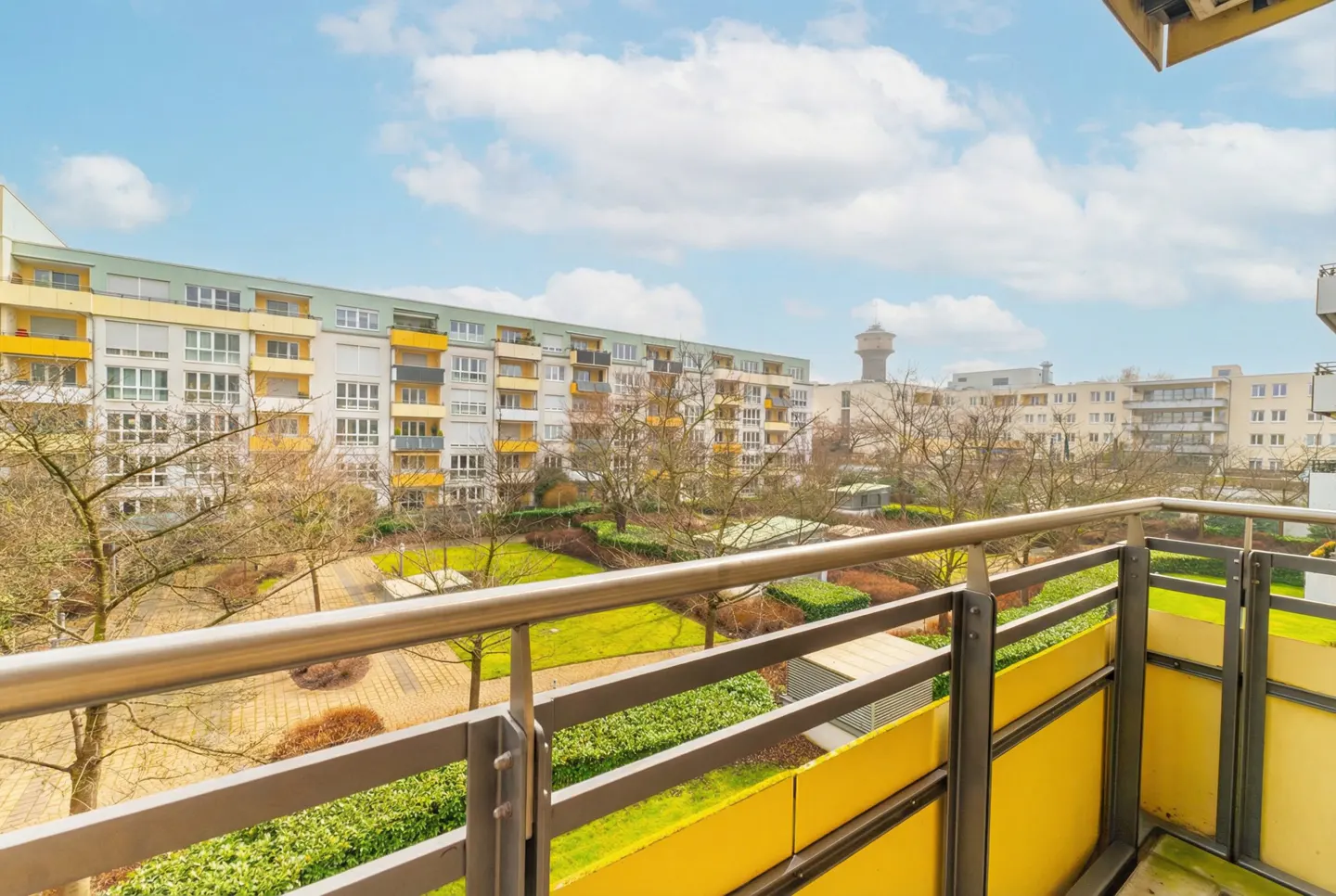 View from a balcony with a metal railing overlooking a green courtyard and apartment buildings under a blue, cloudy sky.