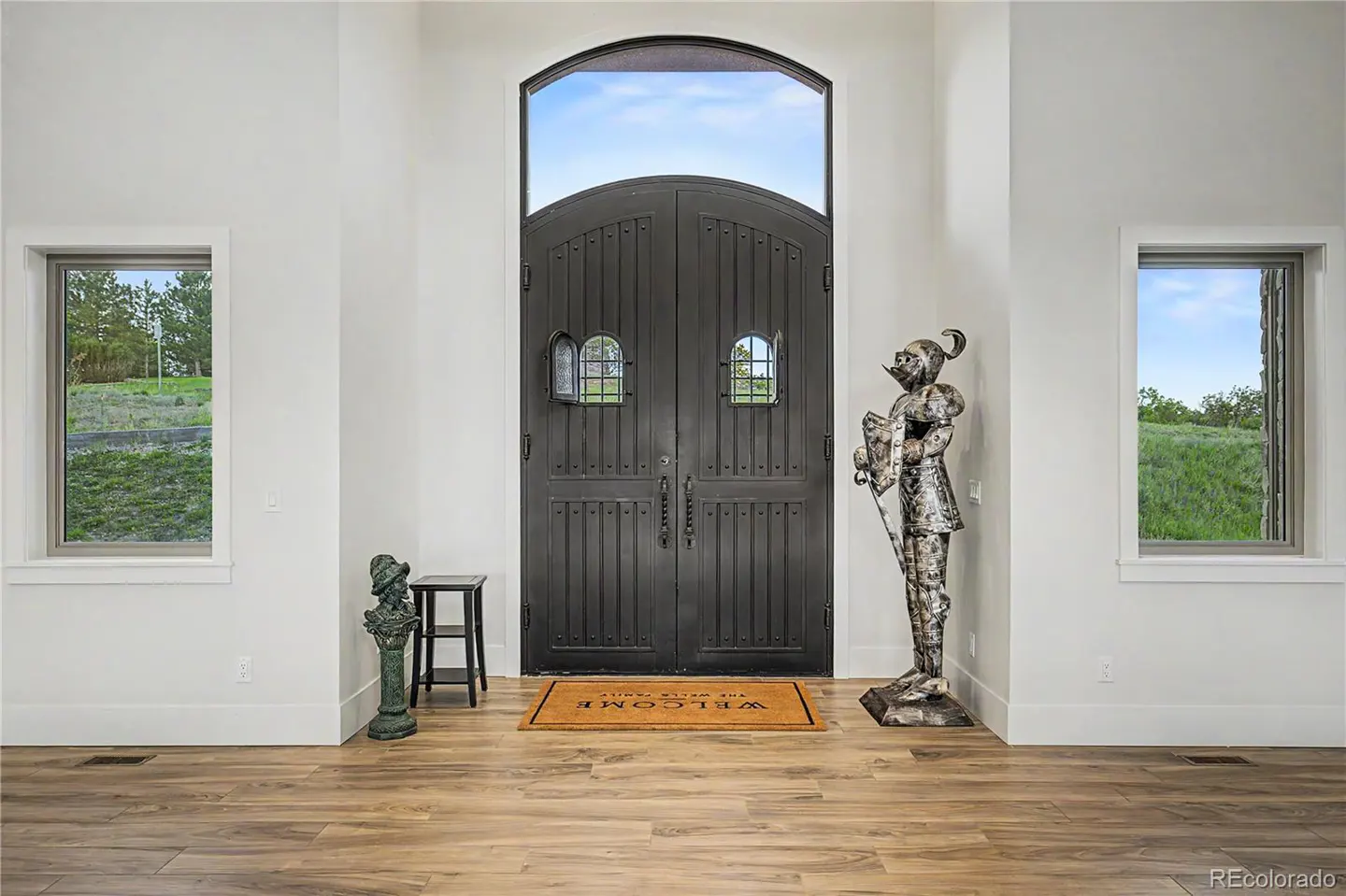 Grand entryway with dark arched double doors, a suit of armor statue, a welcome mat, and two windows overlooking greenery.