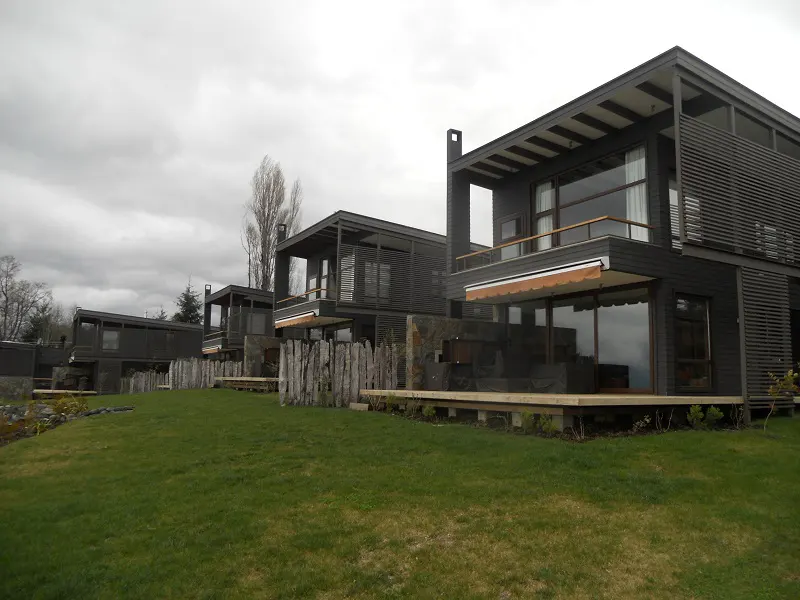 Row of modern, dark wood homes with large windows and decks, separated by a rustic wooden fence, on a green lawn under a cloudy sky.