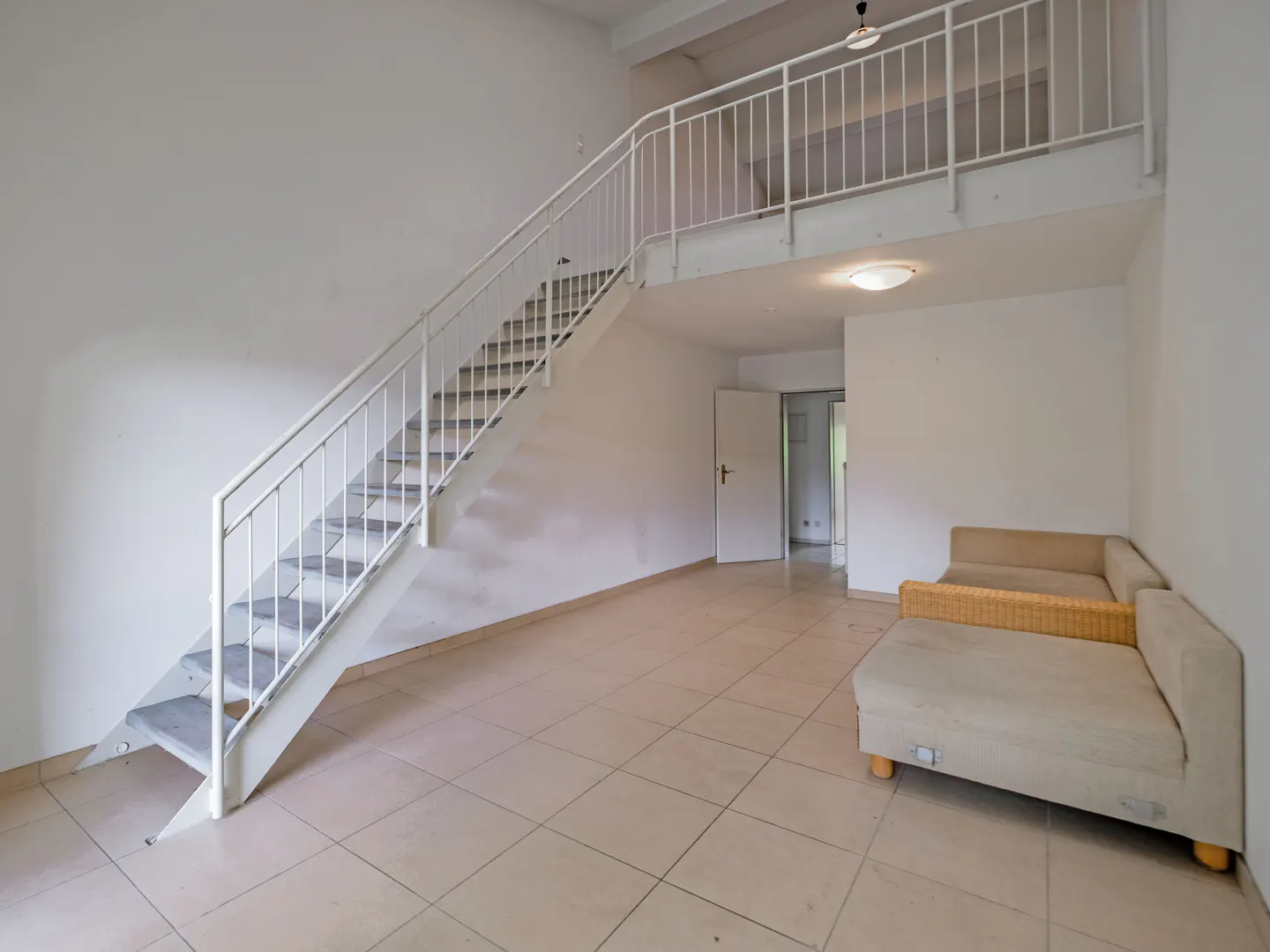 Bright living room with white walls, tiled floors, a beige sofa, and a white staircase leading to an upper loft area.