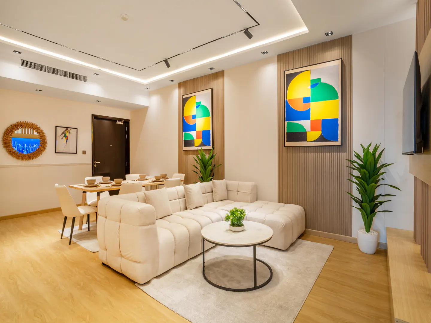 Bright, modern living room with a cream sectional sofa, round coffee table, and dining table set for six. Colorful abstract art adorns the walls.