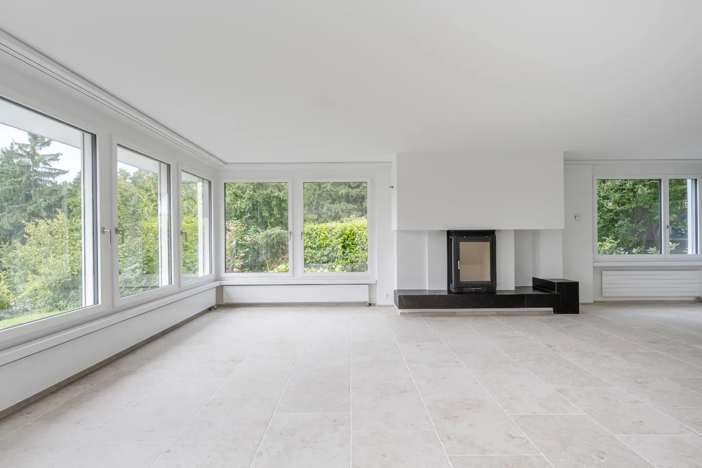 Bright, empty living room with tile floor, white walls, and a black fireplace. Large windows offer a view of lush greenery.