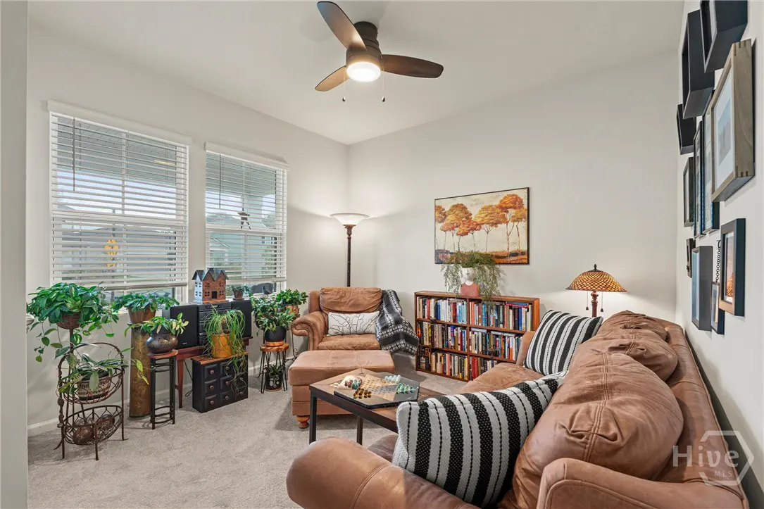 Living room with brown leather sofa, chair, and ottoman. Bookshelf, plants, and artwork decorate the space. A ceiling fan is overhead.