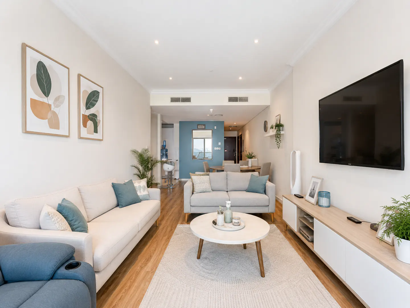 Living room with white sofas, teal pillows, round coffee table, TV, and abstract art. Neutral tones and wood floors.